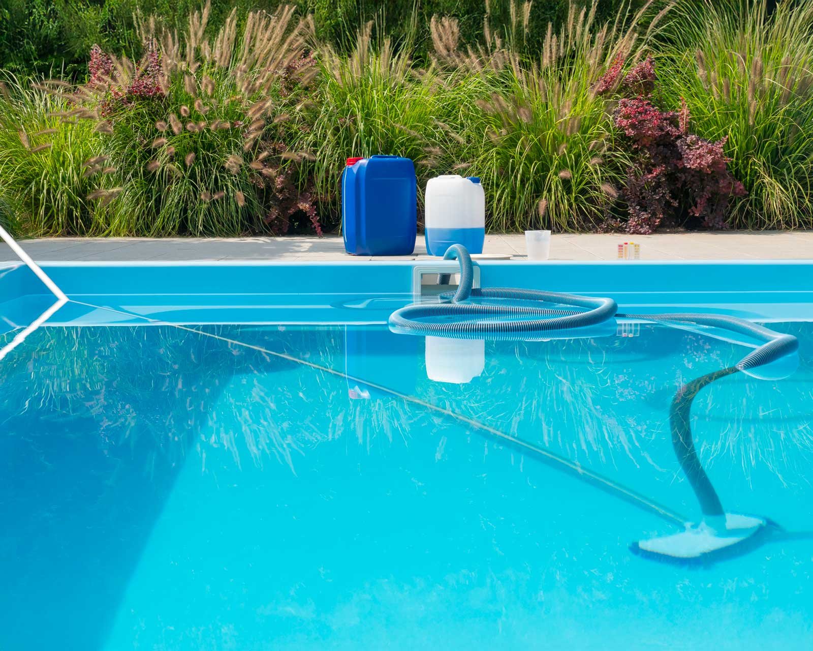 Tuyau et brosse de nettoyage dans une piscine, bidons de traitement et bandelettes de test posés sur la margelle
