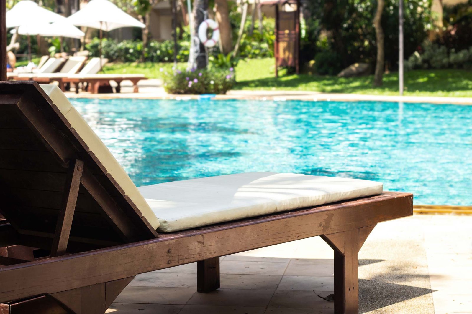 Transat en bois au premier plan au bord d’une grande piscine, parasols en arrière-plan
